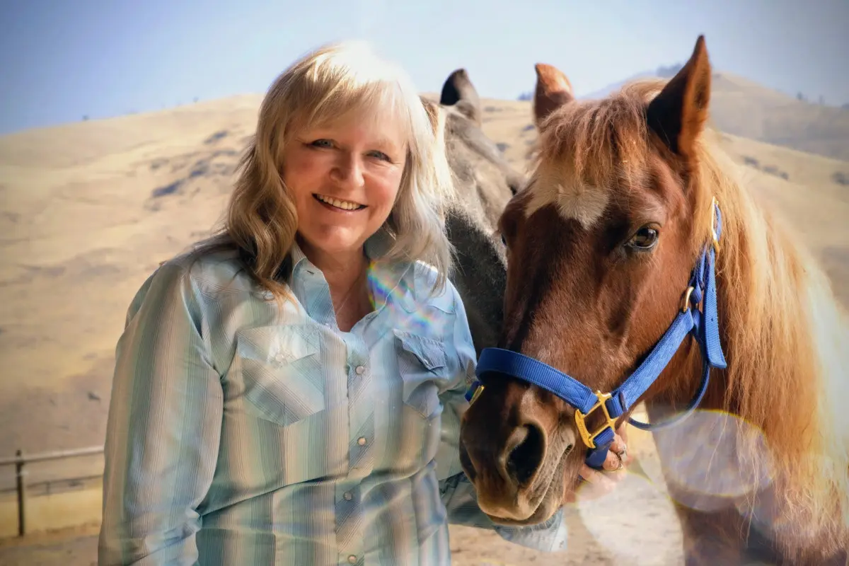 A smiling woman stands next to a horse in a rural landscape.