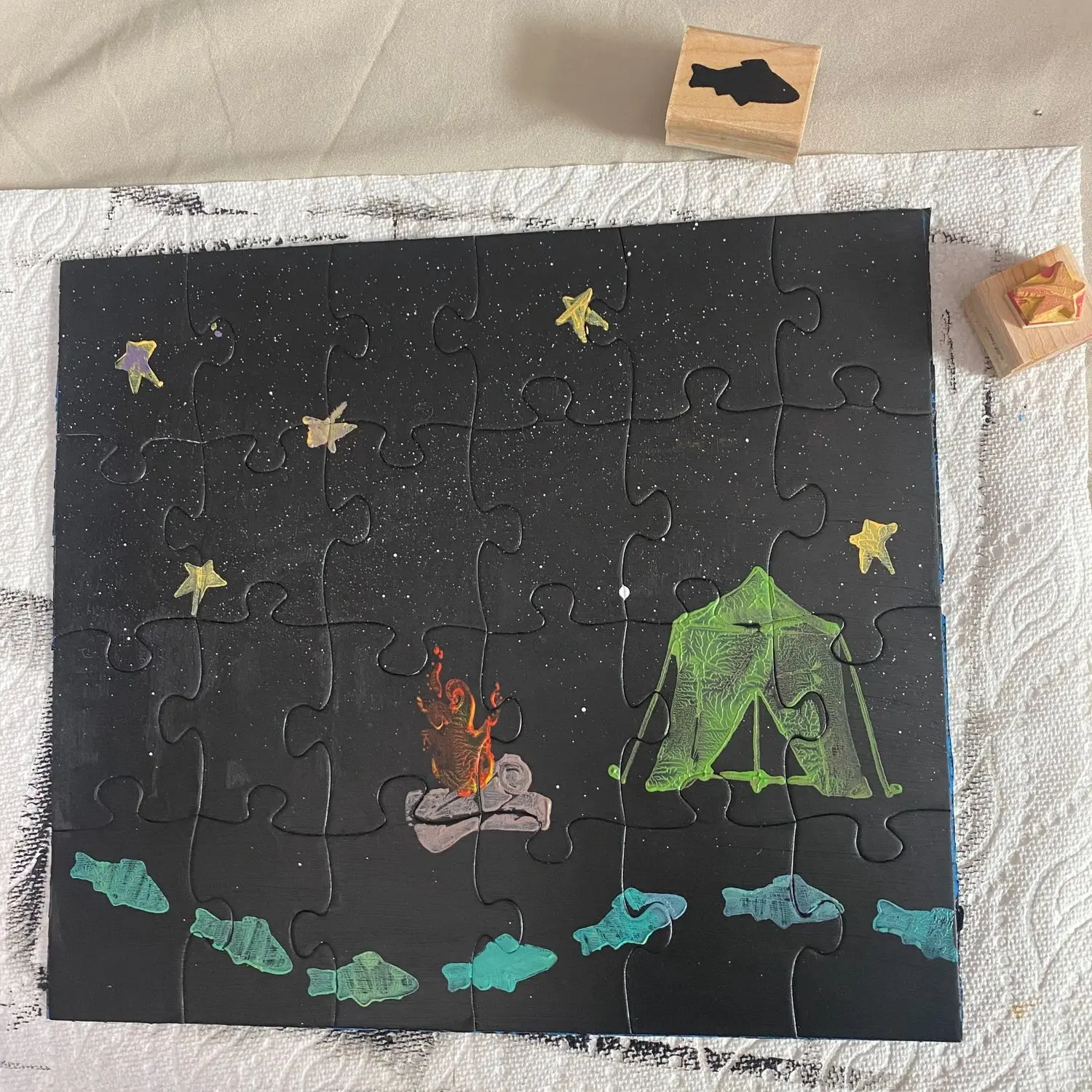 A completed puzzle depicts a nighttime camping scene with a tent, fire, and fish motifs.