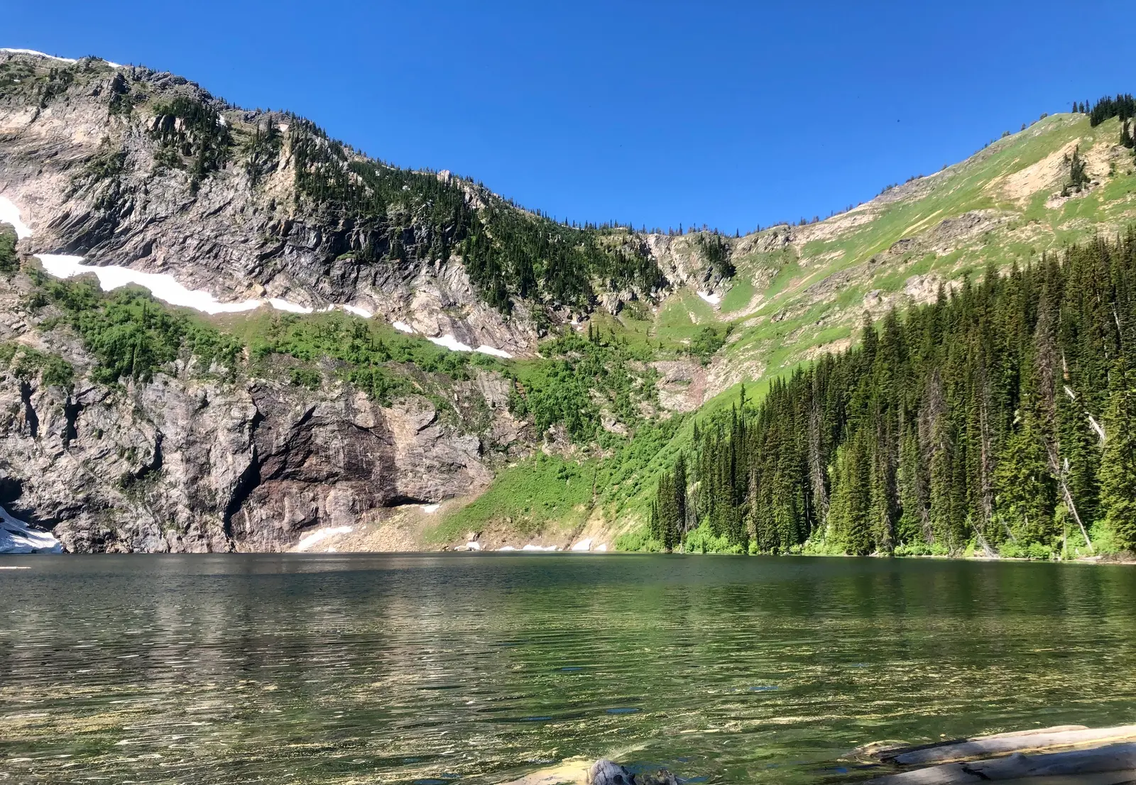 A mountain lake reflects high peaks with patches of snow and dense evergreen forests.
