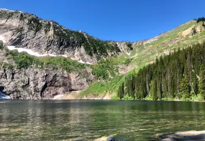 A mountain lake reflects high peaks with patches of snow and dense evergreen forests.