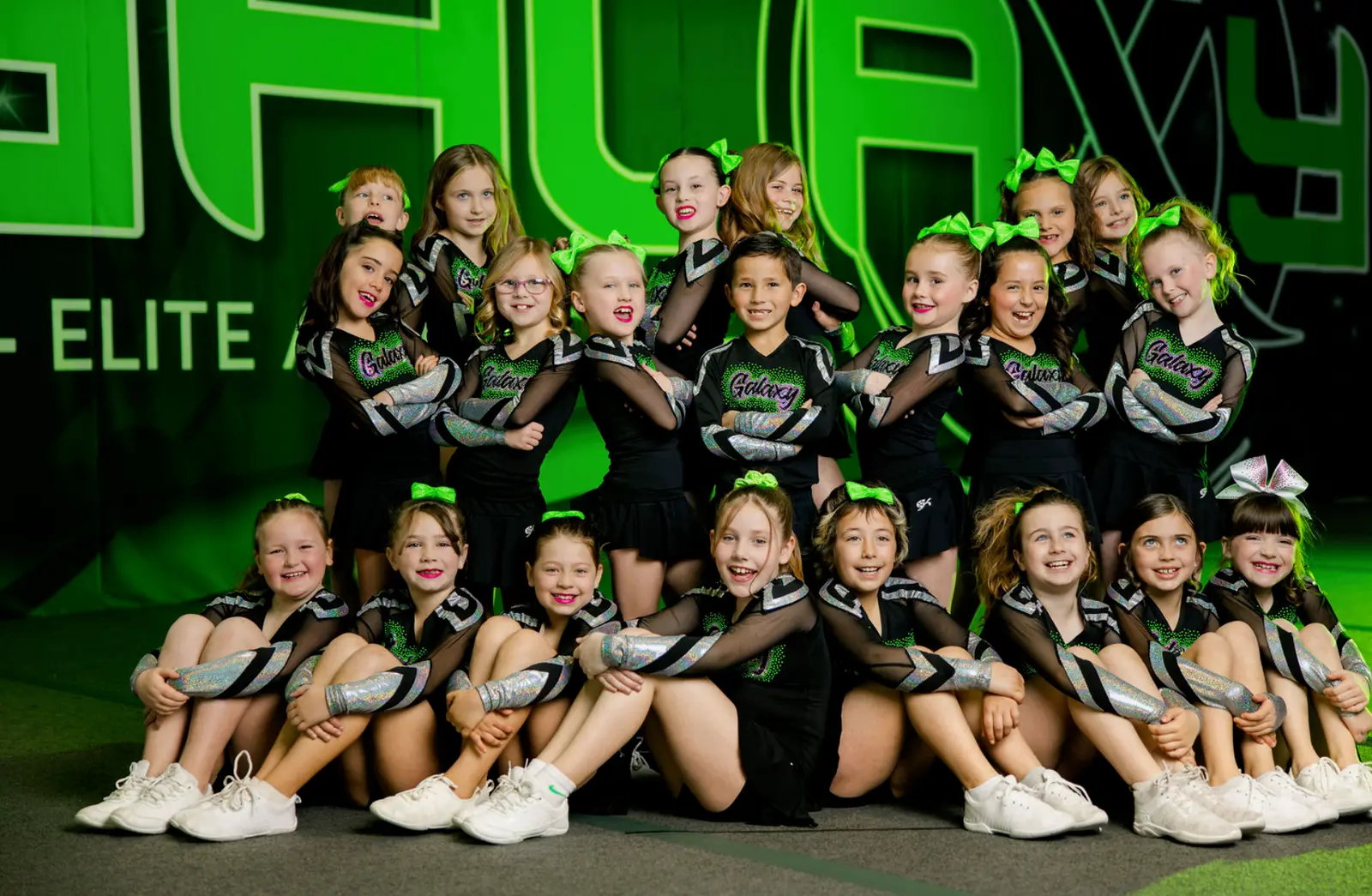 A large group of children in matching cheerleading uniforms pose for a photo in front of a branded backdrop.