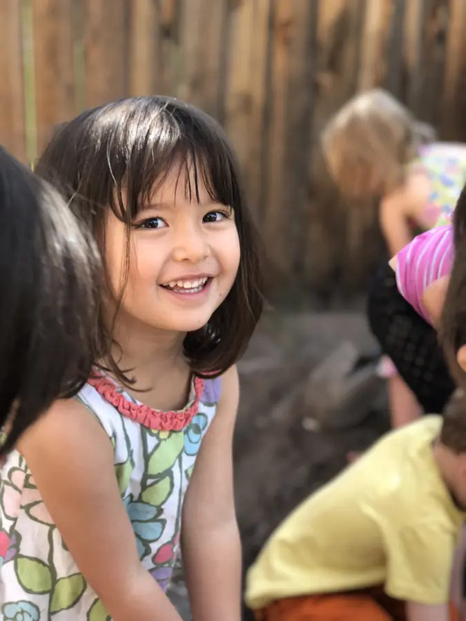 A smiling child looks at the camera while playing outdoors with other children.