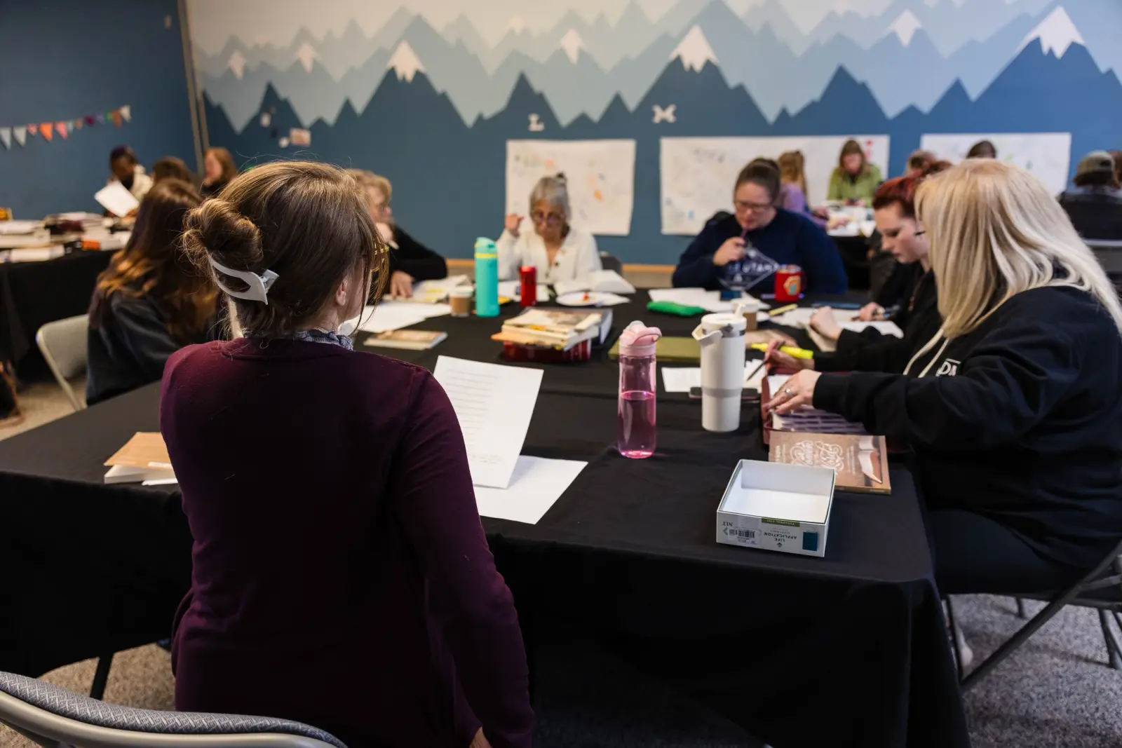 A group of adults are seated around a table for a meeting or study session in a room with mountain-themed wall art.