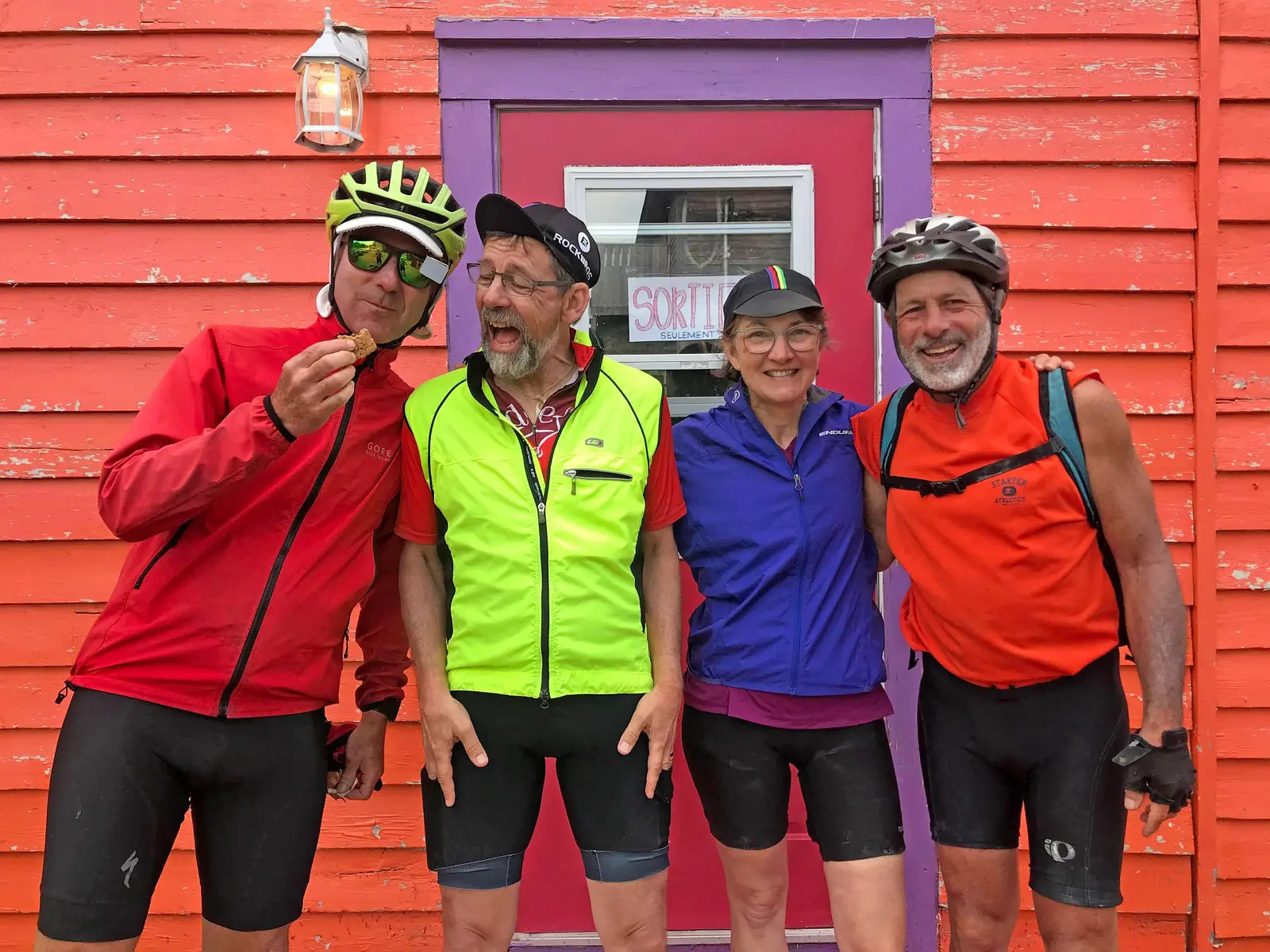 A group of four cyclists in athletic gear stand together against a bright red wall.