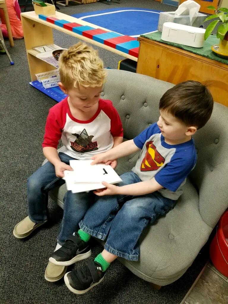 Two young boys sit together on a small gray couch in a classroom holding and looking at flashcards.