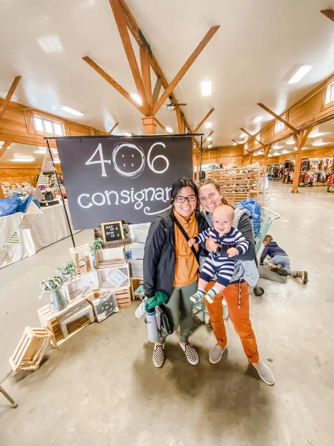 Three people, two women and a baby, smiling and posing together inside a store with wooden beams. A black sign with white text reads '406 consignment' behind them. The store has various items displayed on tables and shelves.