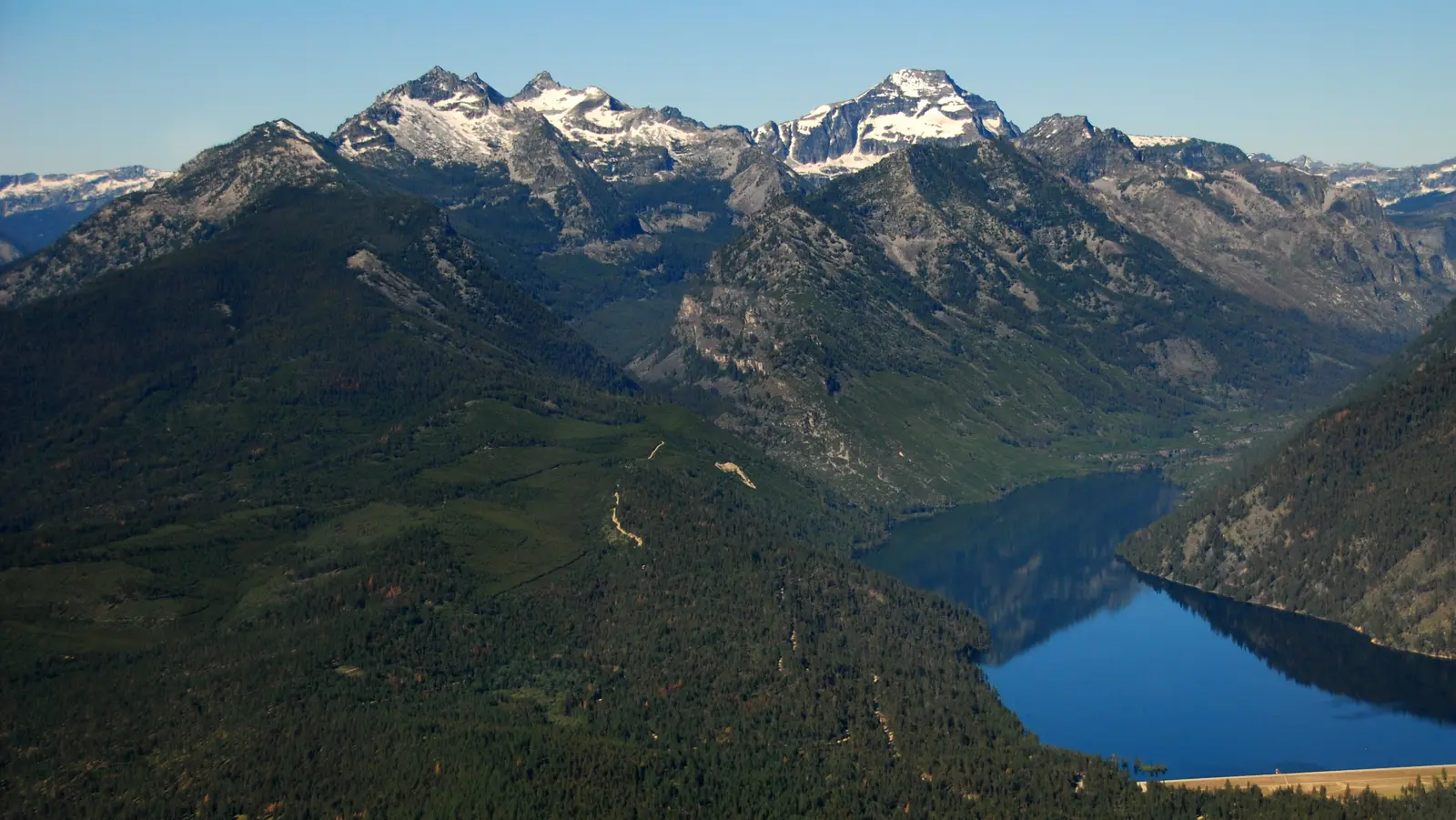 An aerial view shows snow-capped mountains overlooking a lake and forested terrain.