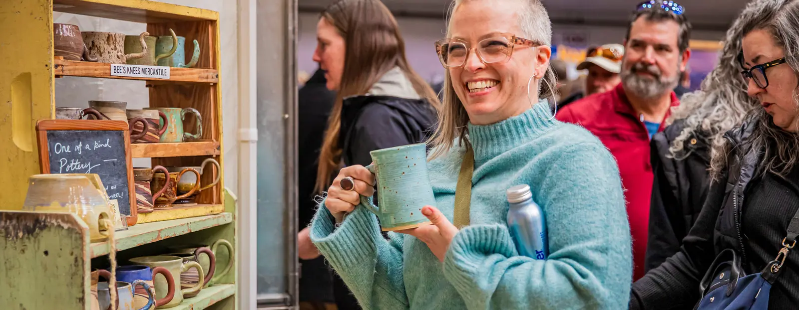 A smiling woman holds a handmade pottery mug while browsing at a craft stall with other shoppers in the background.