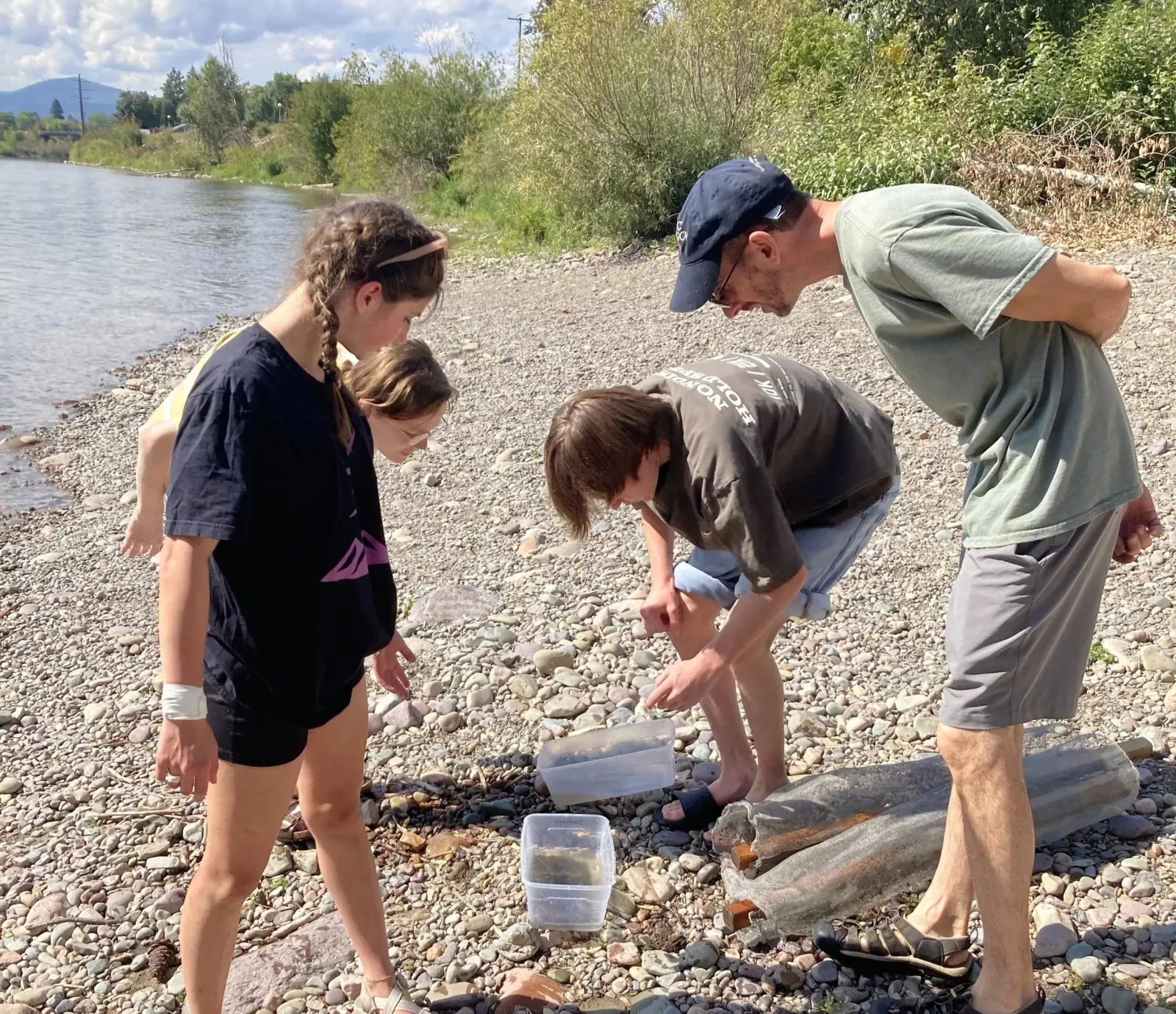An instructor and three youths examine samples in plastic containers by a riverbank.