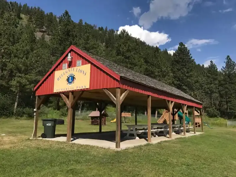 A wooden pavilion with a red roof stands in a park with playground equipment in the background.
