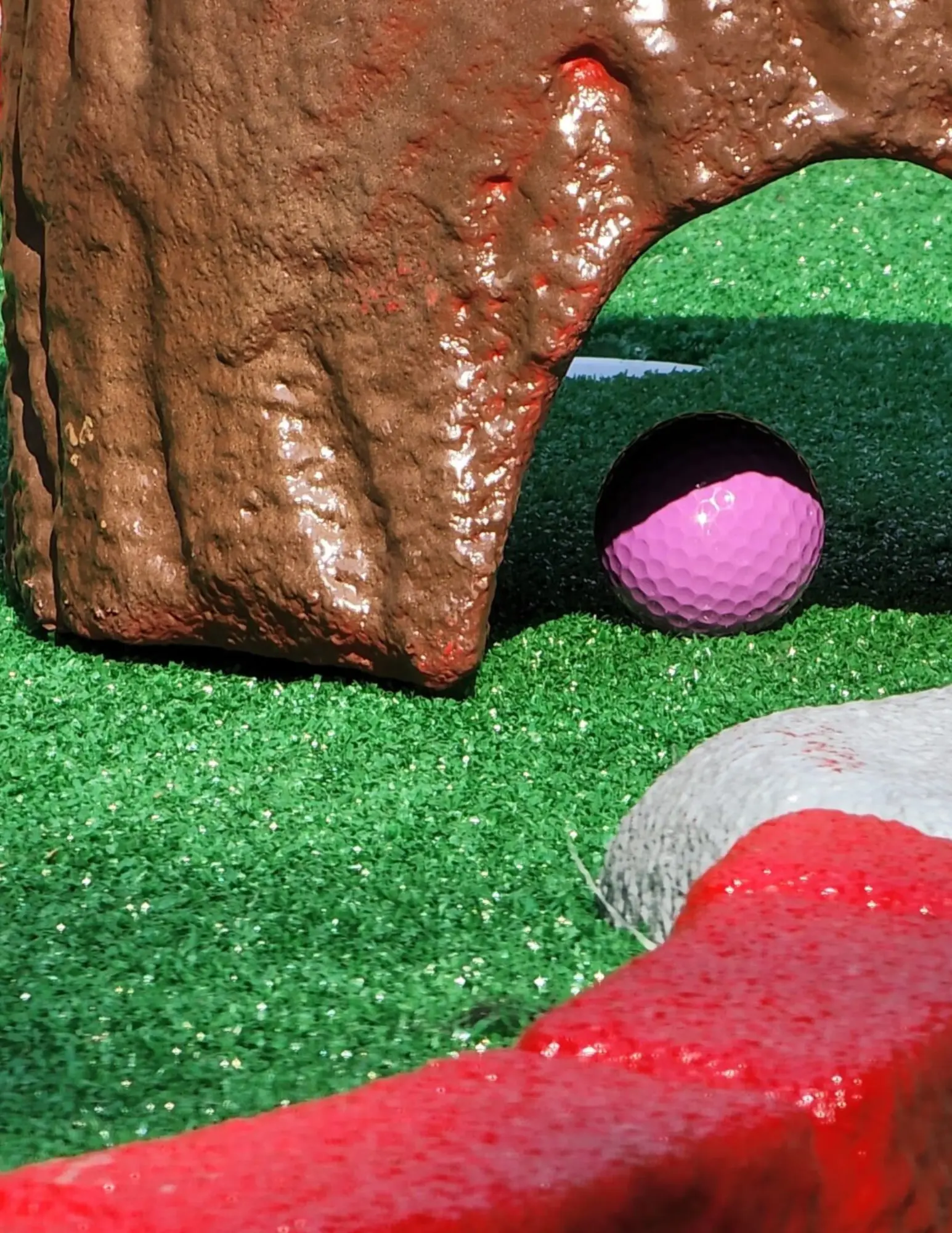 A purple golf ball sits on a miniature golf course near a decorative log structure.