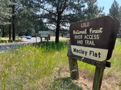 Photo from fs.usda.gov A sign for a river access and trail area stands in a grassy clearing with a parking area visible in the background.