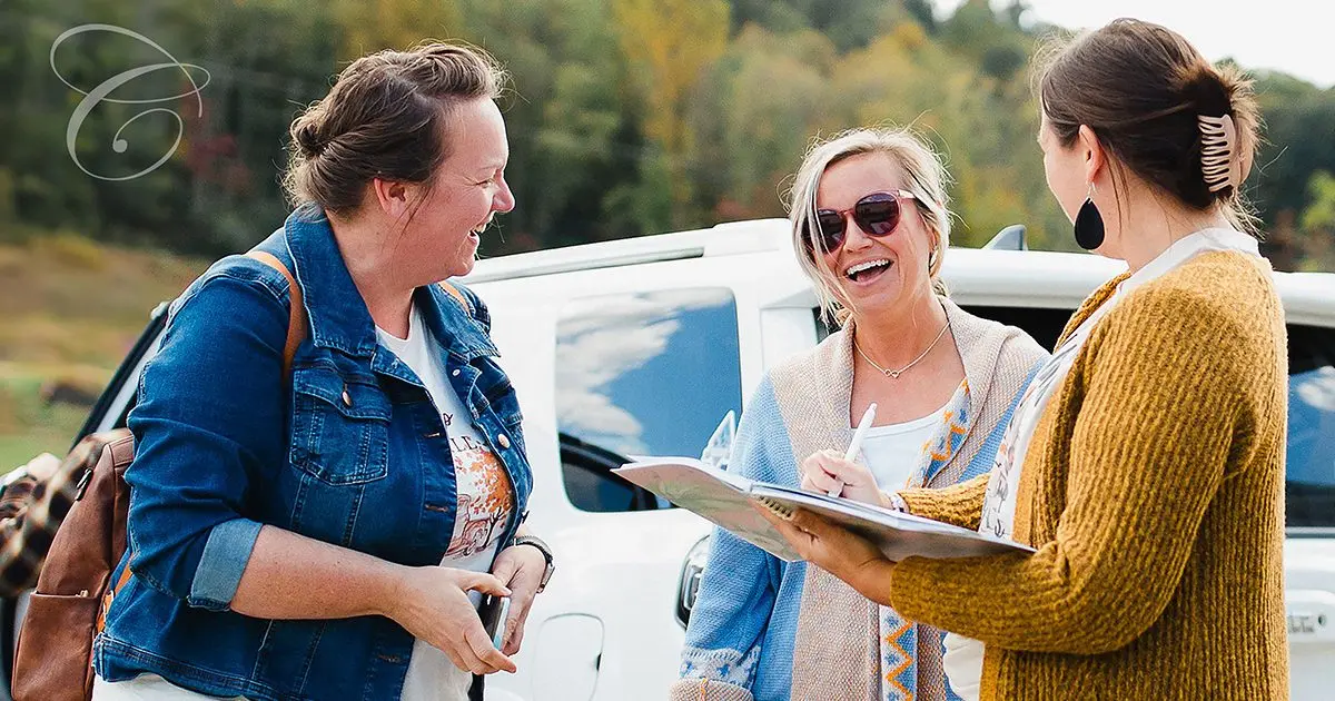Three adult women are laughing and talking outdoors near a white vehicle.