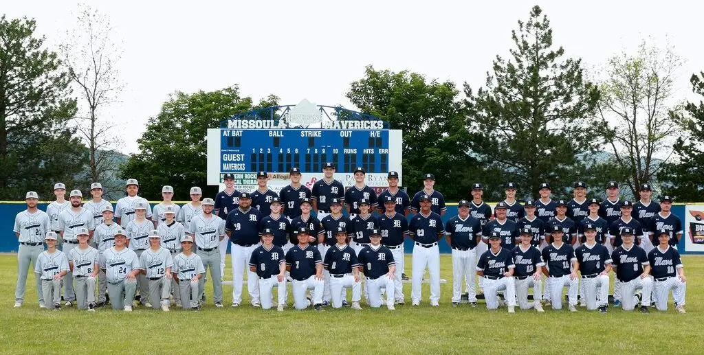 A large baseball team poses for a group photo on a grassy field in front of a scoreboard.