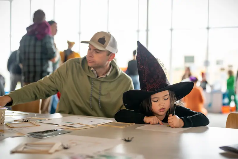 A child wearing a witch hat draws at a table while a man sits beside them.