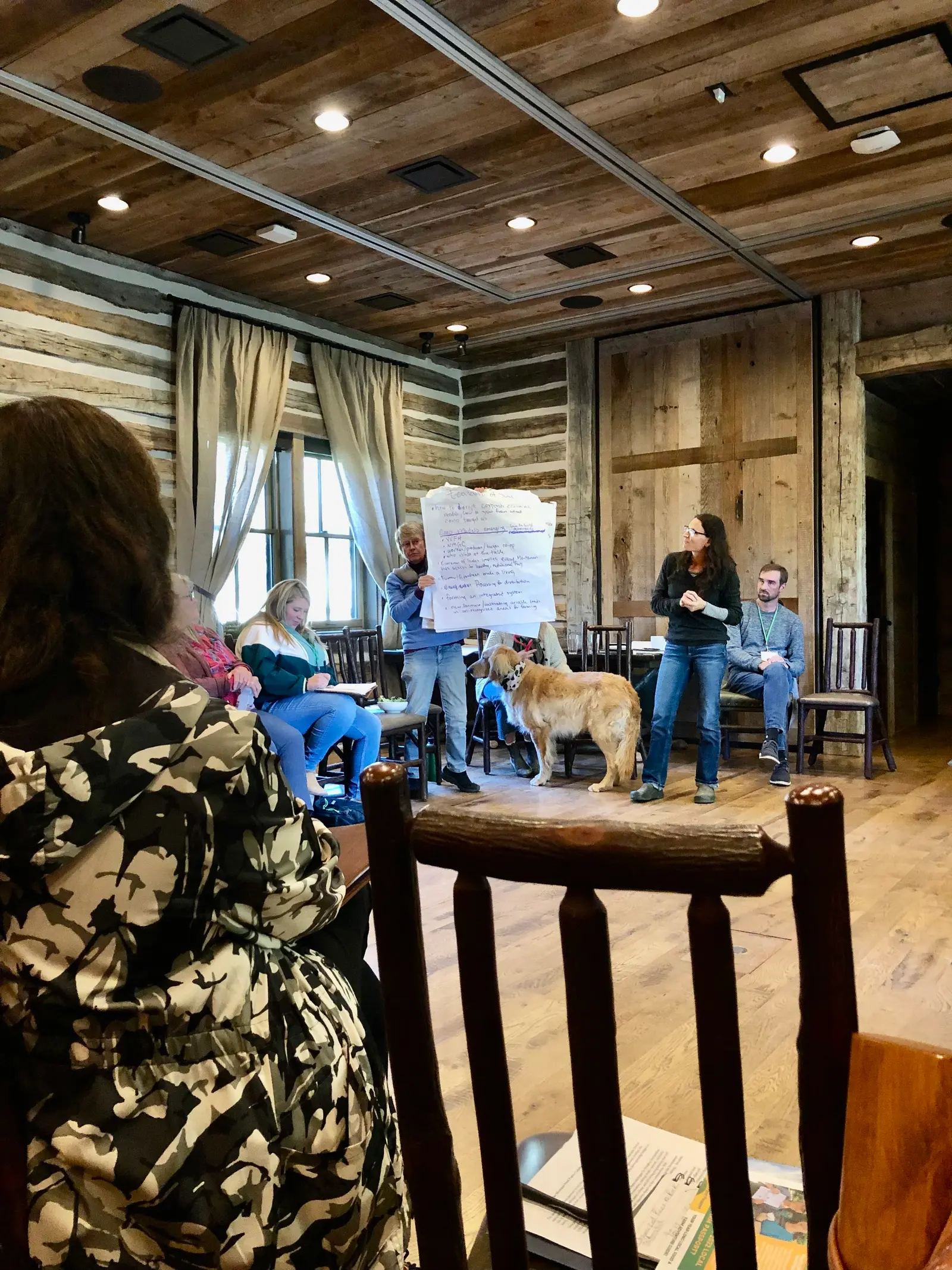 A group of adults sits in a rustic room while a woman holds up a poster for a presentation with a golden retriever nearby.