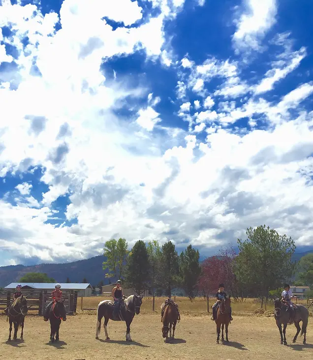 A group of riders on horseback stand in a dirt arena under a cloudy blue sky.