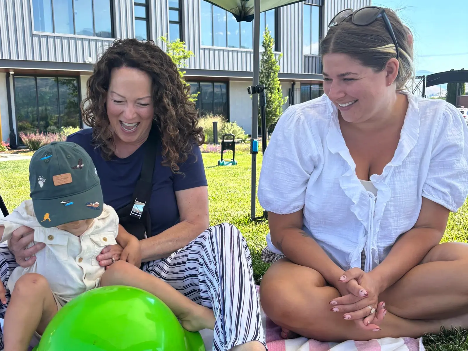 Two women sit on the grass outdoors with a young child.