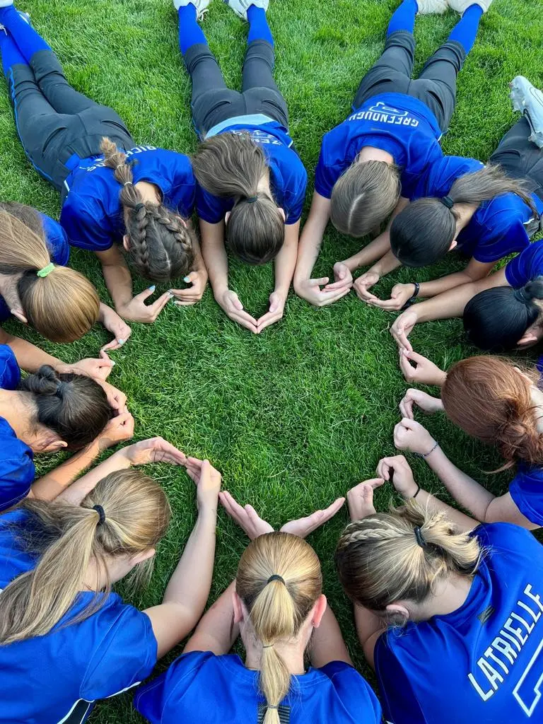 A group of players in blue jerseys lie on the grass in a circle with their hands touching to form a shape.