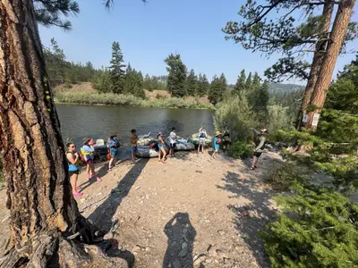 A group of people are gathered on a riverbank with rafts, with one individual appearing to provide instructions.