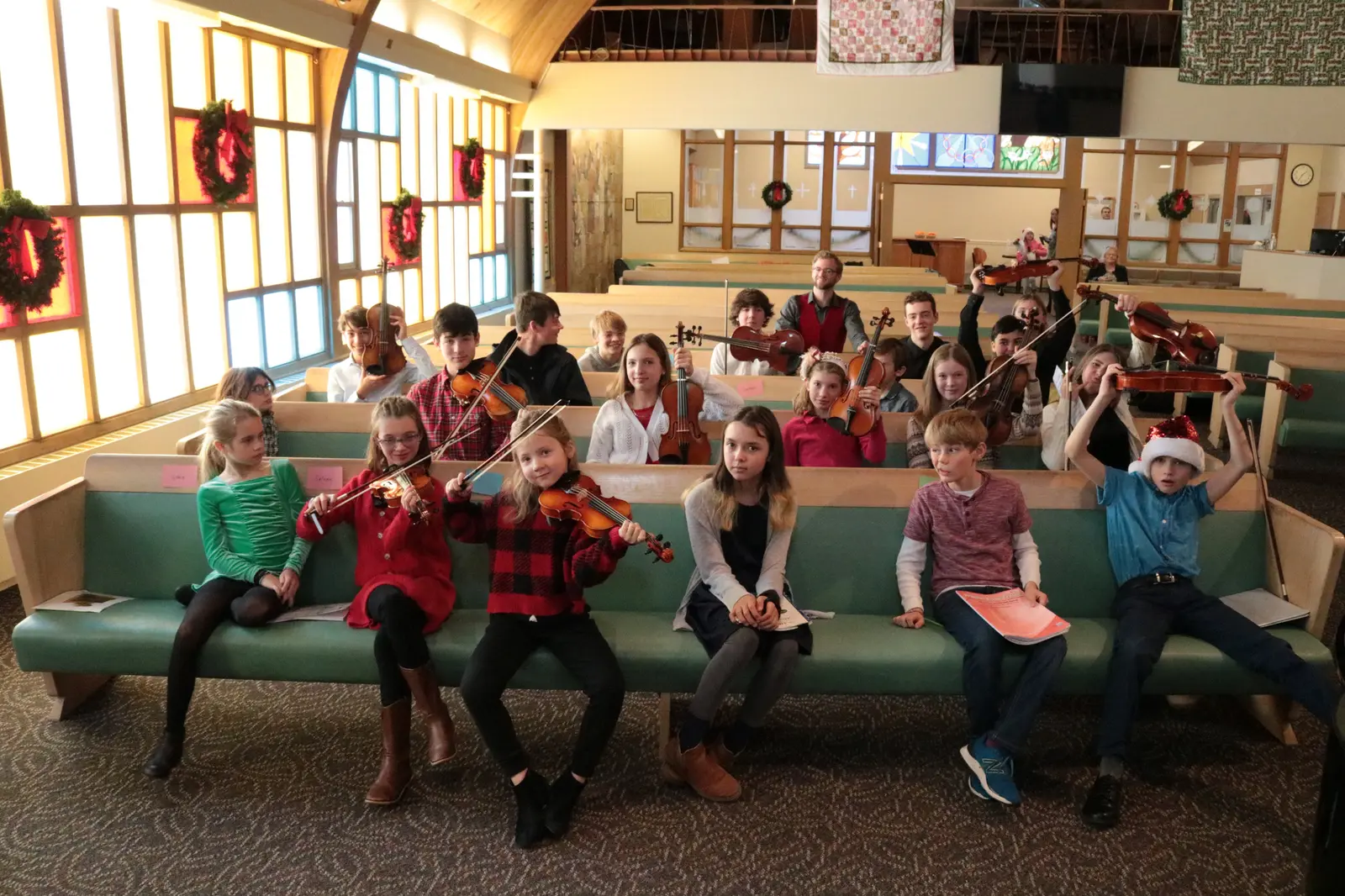 A group of children and an instructor holding violins sit on pews in a church hall.