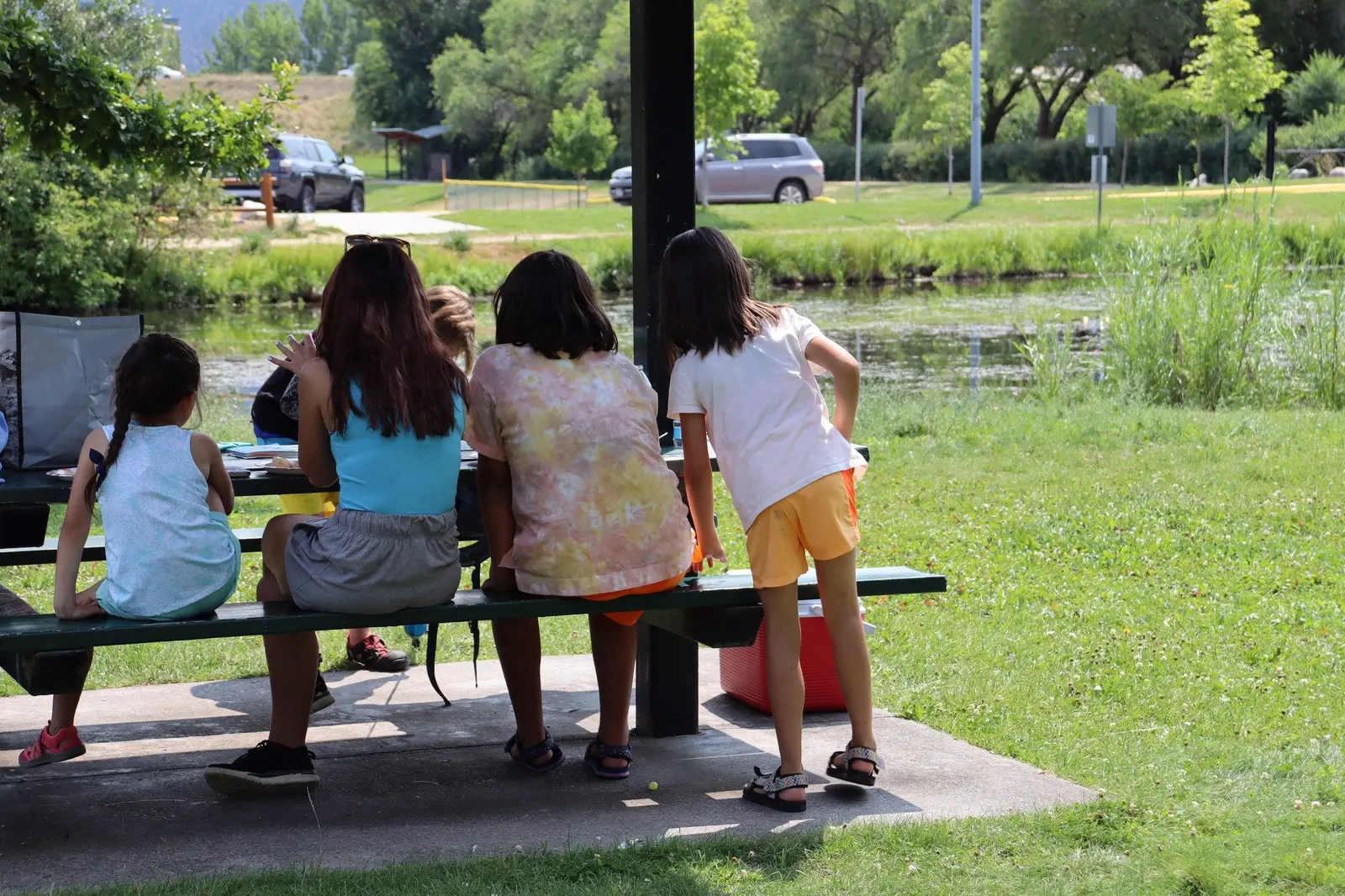 A group of children sits at a picnic table outdoors near a pond.