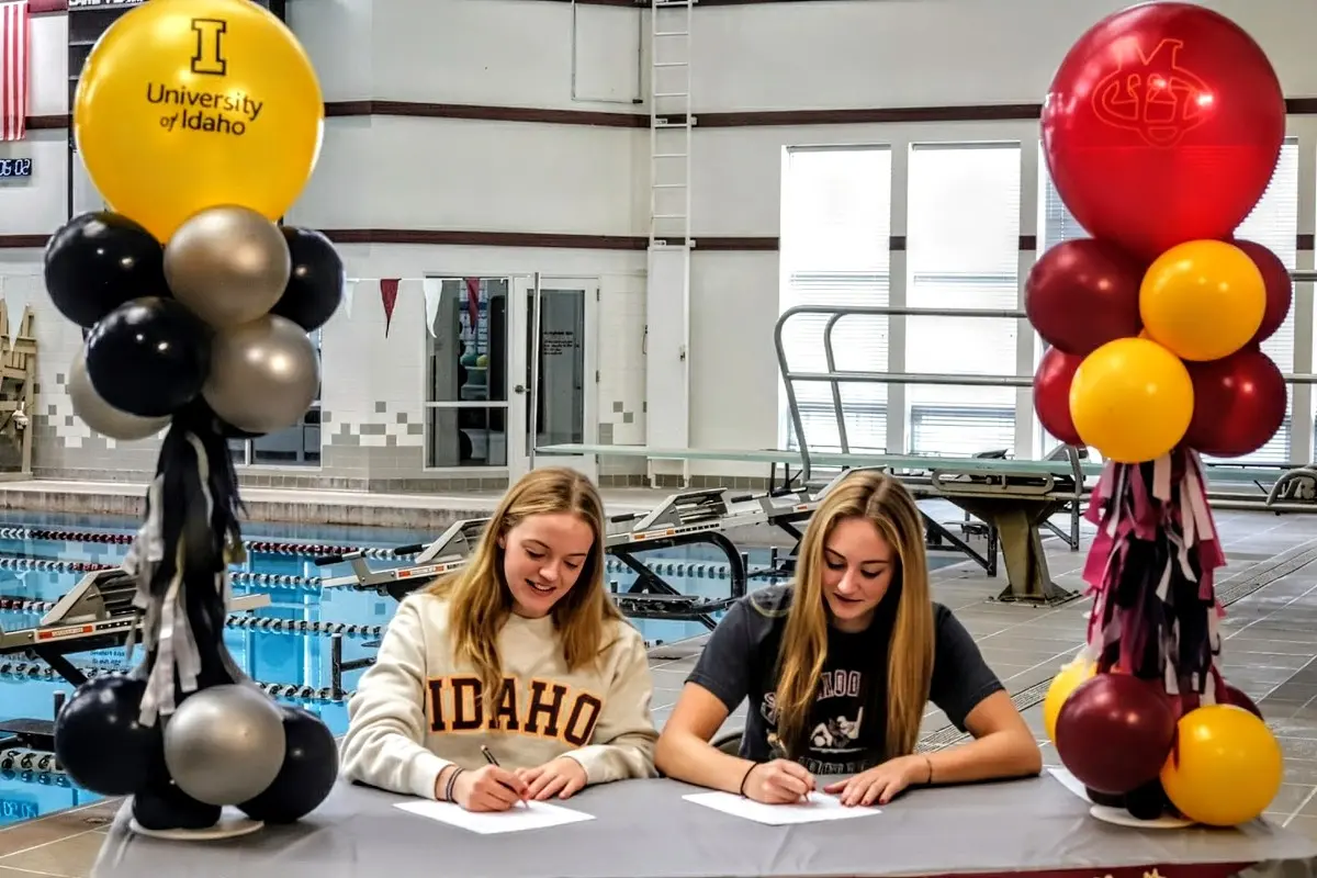 Two students sit at a table signing papers in an indoor swimming pool facility decorated with balloons.