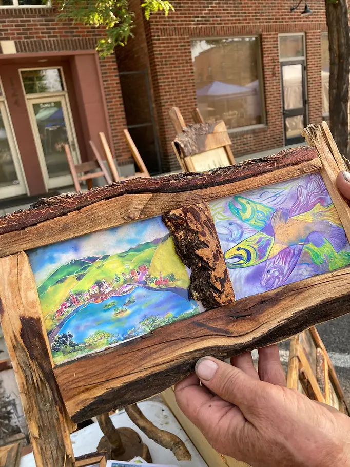 A person holds a rustic wooden frame containing two panels depicting a landscape and stylized fish.