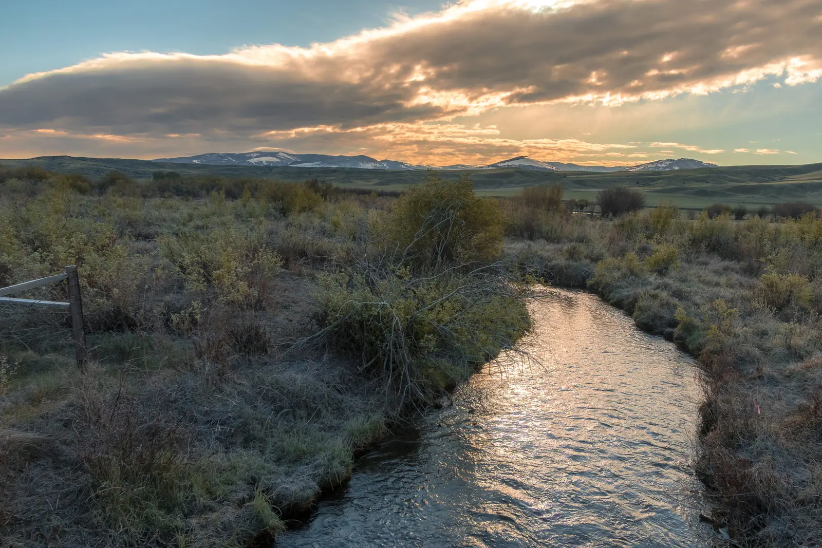 A stream flows through a grassy meadow under a cloudy sunset sky with mountains in the background.