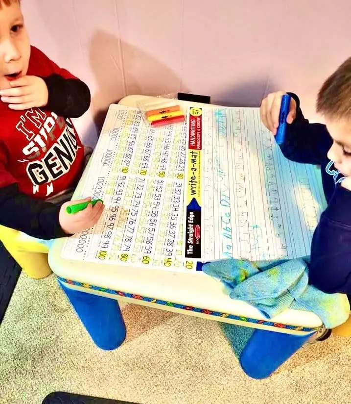 Two children sit at a small table while practicing handwriting and math with educational tools.