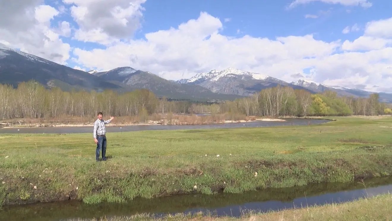 A man stands in a grassy field with a river and mountains in the background.