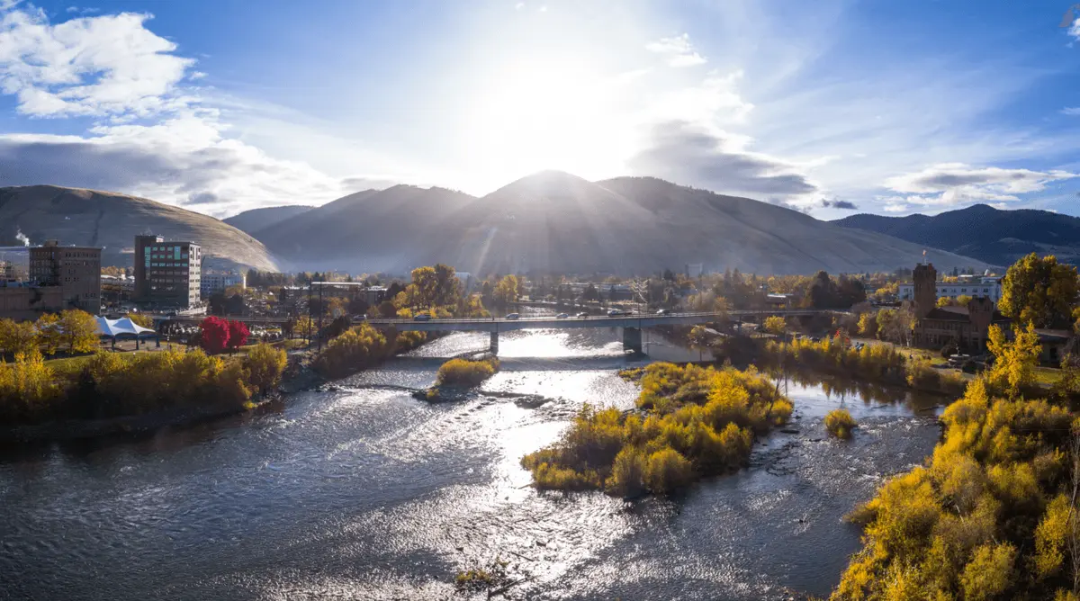 A river flows through a town with mountains in the background under a bright sun.
