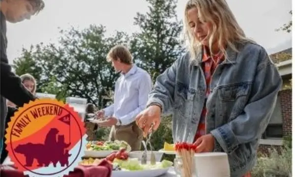 Young adults serve themselves food at an outdoor event with a decorative Family Weekend logo overlay.