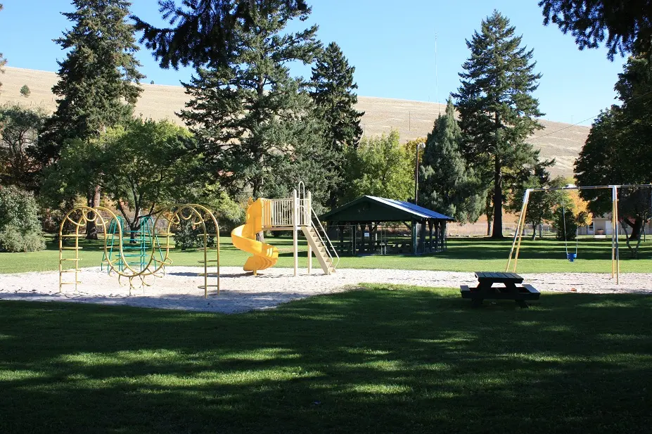 A public playground area features climbing equipment, a yellow slide, and a picnic table in a green park.