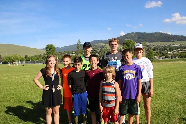 A group of children and an instructor pose for a photo on a grassy field with mountains in the background.