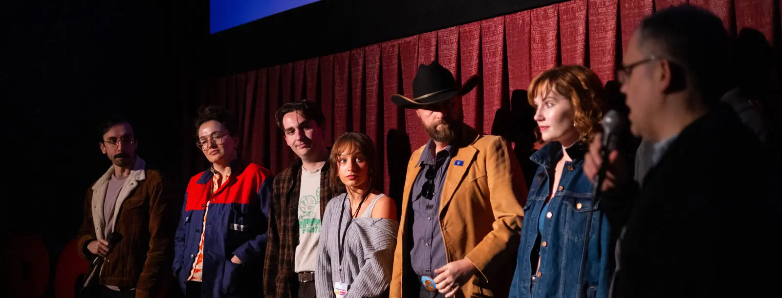A group of adults stands on a stage in front of a red curtain as one speaks into a microphone.