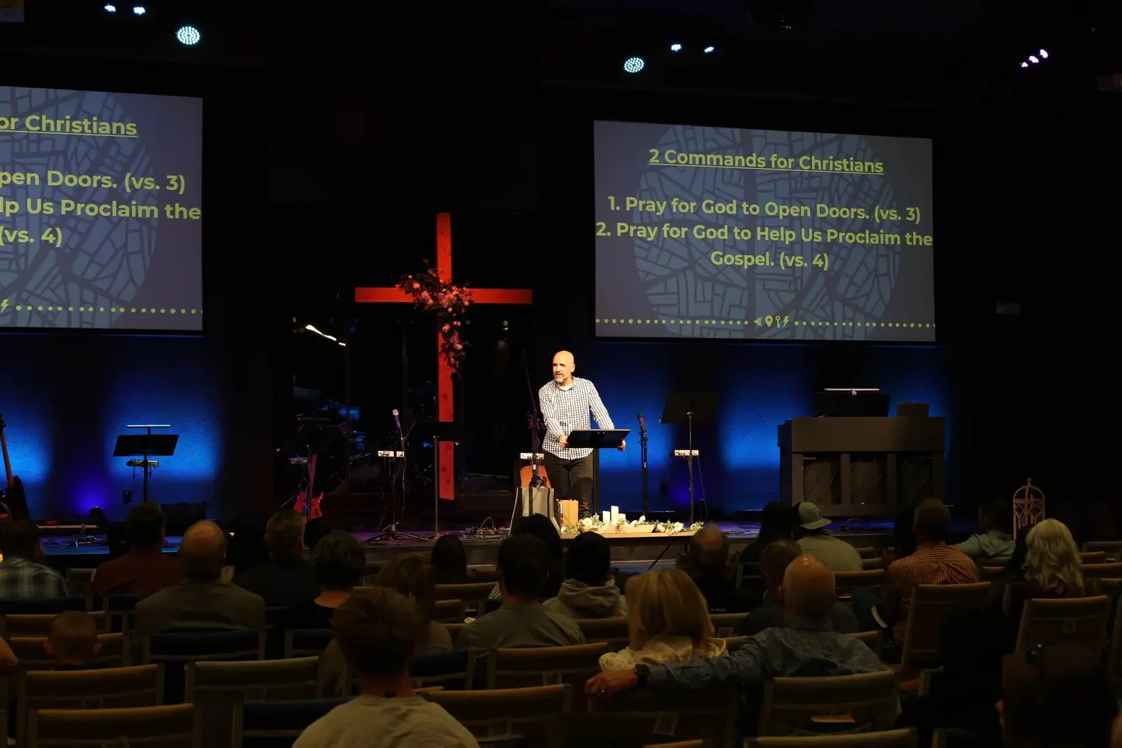 A man stands at a lectern on a stage in front of a seated audience, with projected text visible on screens behind him.