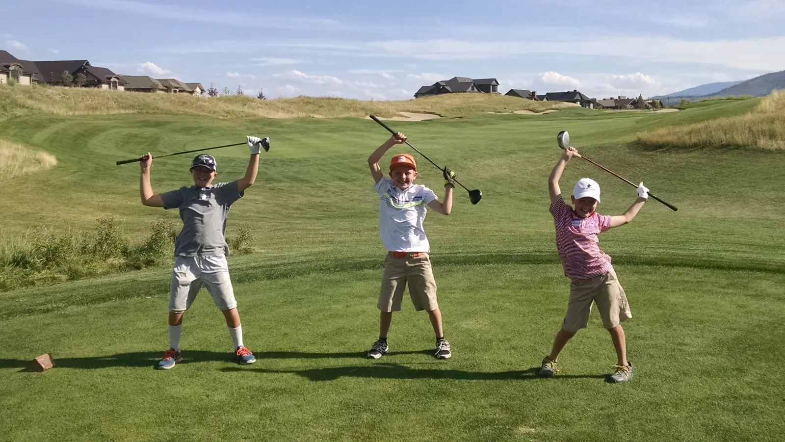 Three children stand on a golf course with their golf clubs raised in the air.