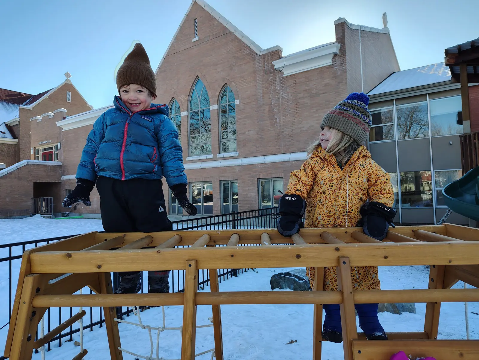 Two children play on a wooden climbing structure outside in the snow.