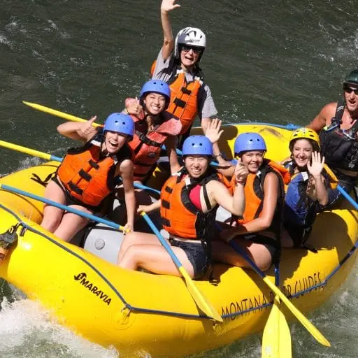 A group of people are smiling and waving while white-water rafting in a yellow raft.