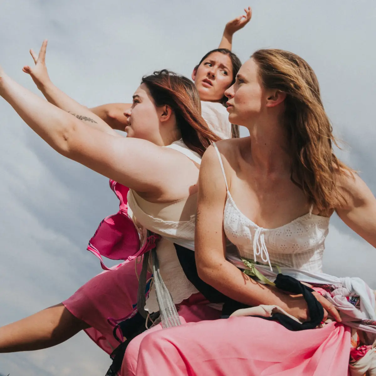 Three women pose together against a cloudy sky with raised arms.