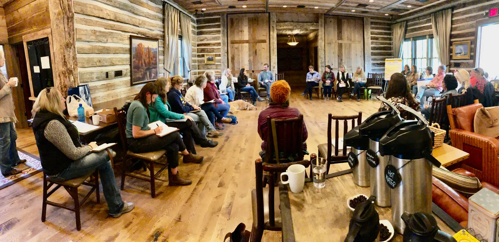 A group of adults sits in a circle inside a rustic room for a meeting.