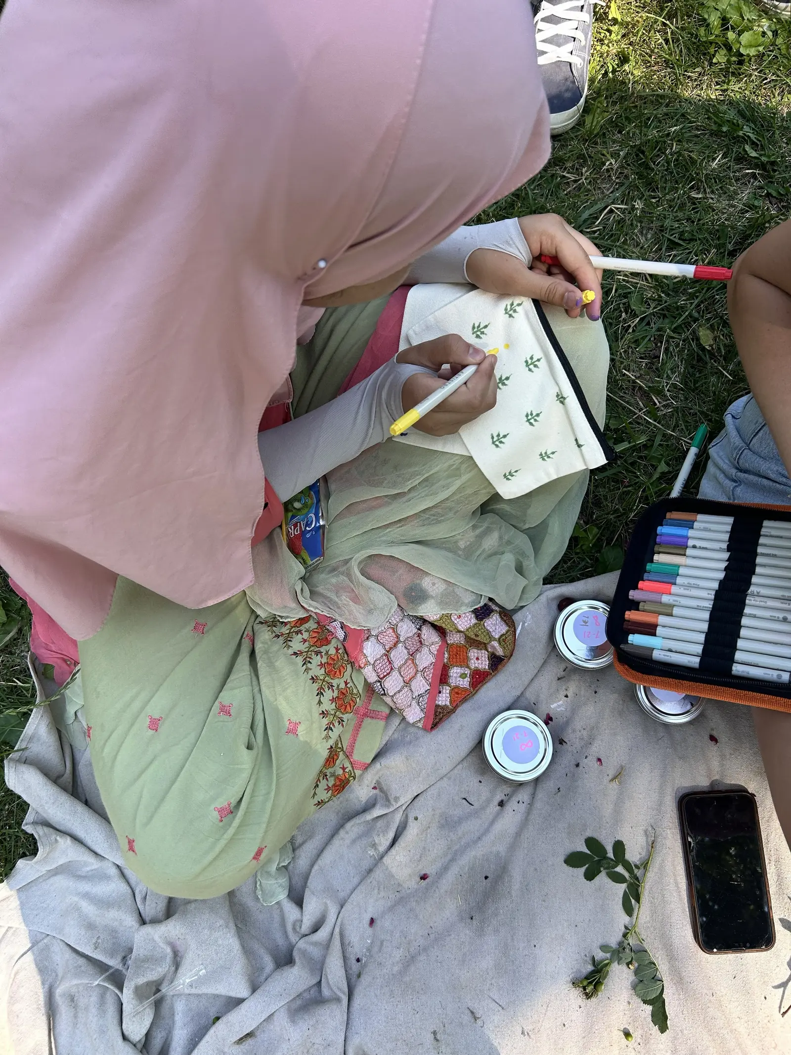 A person wearing a headscarf sits on the ground outdoors, drawing on a small fabric pouch with markers.