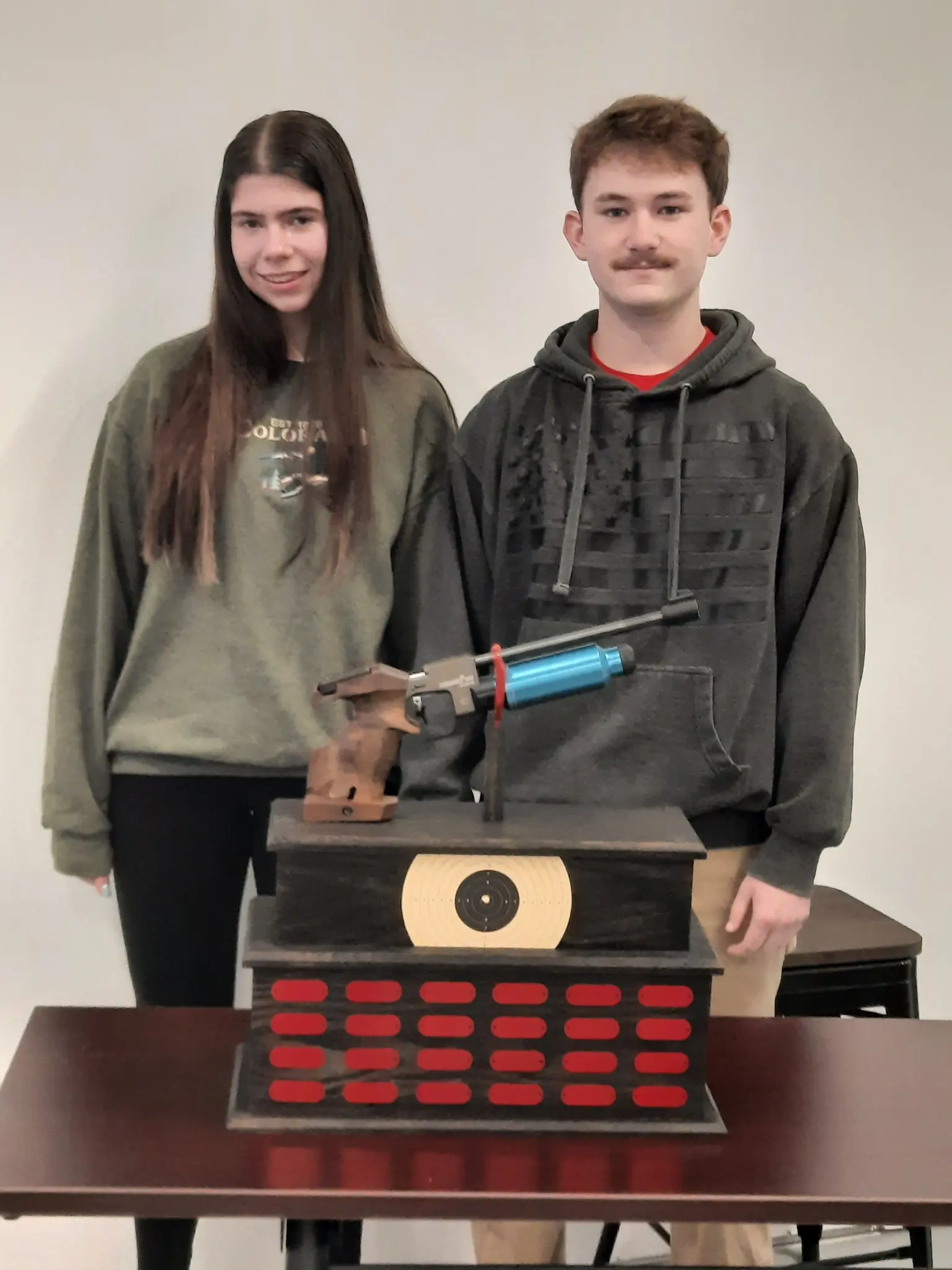 Two teenagers stand behind a wooden trophy featuring a target face and a competition pistol.