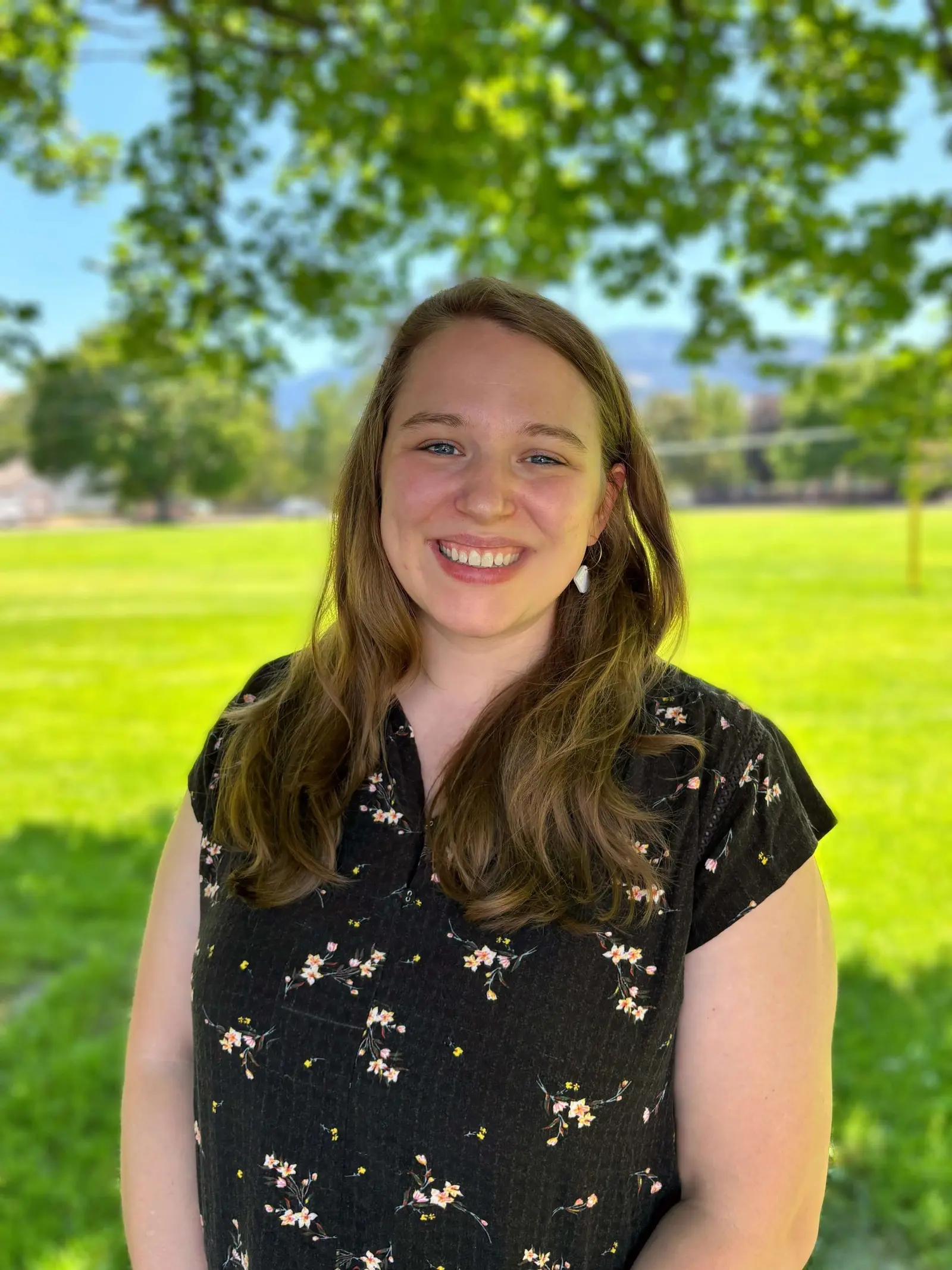A smiling woman stands in a grassy park on a sunny day.