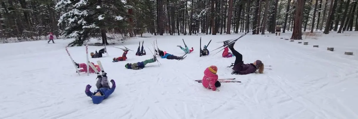 A group of children are lying in the snow with their skis on in a forest clearing.