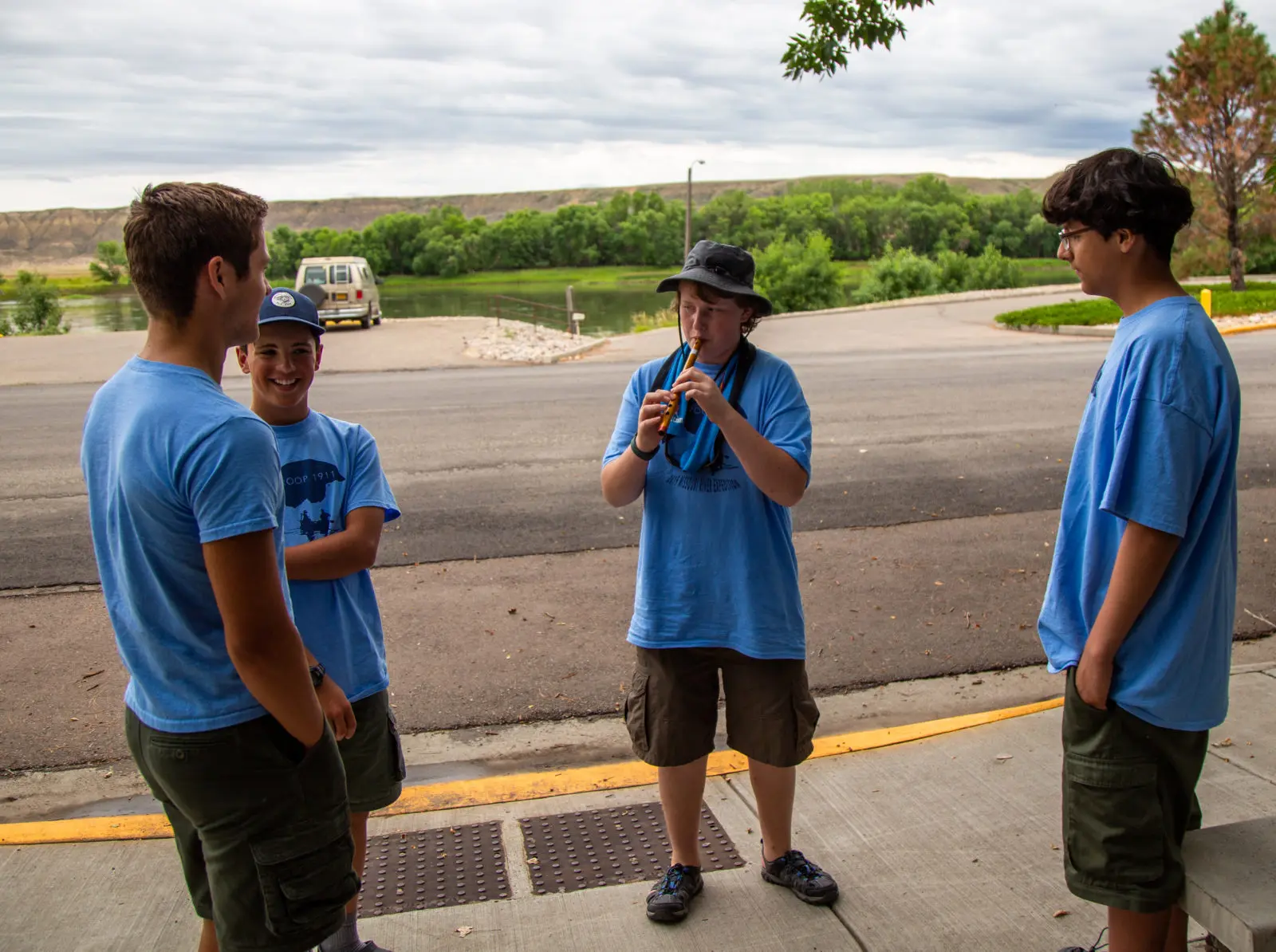 Four young people in light blue t-shirts gather outdoors, with one playing a flute-like instrument.