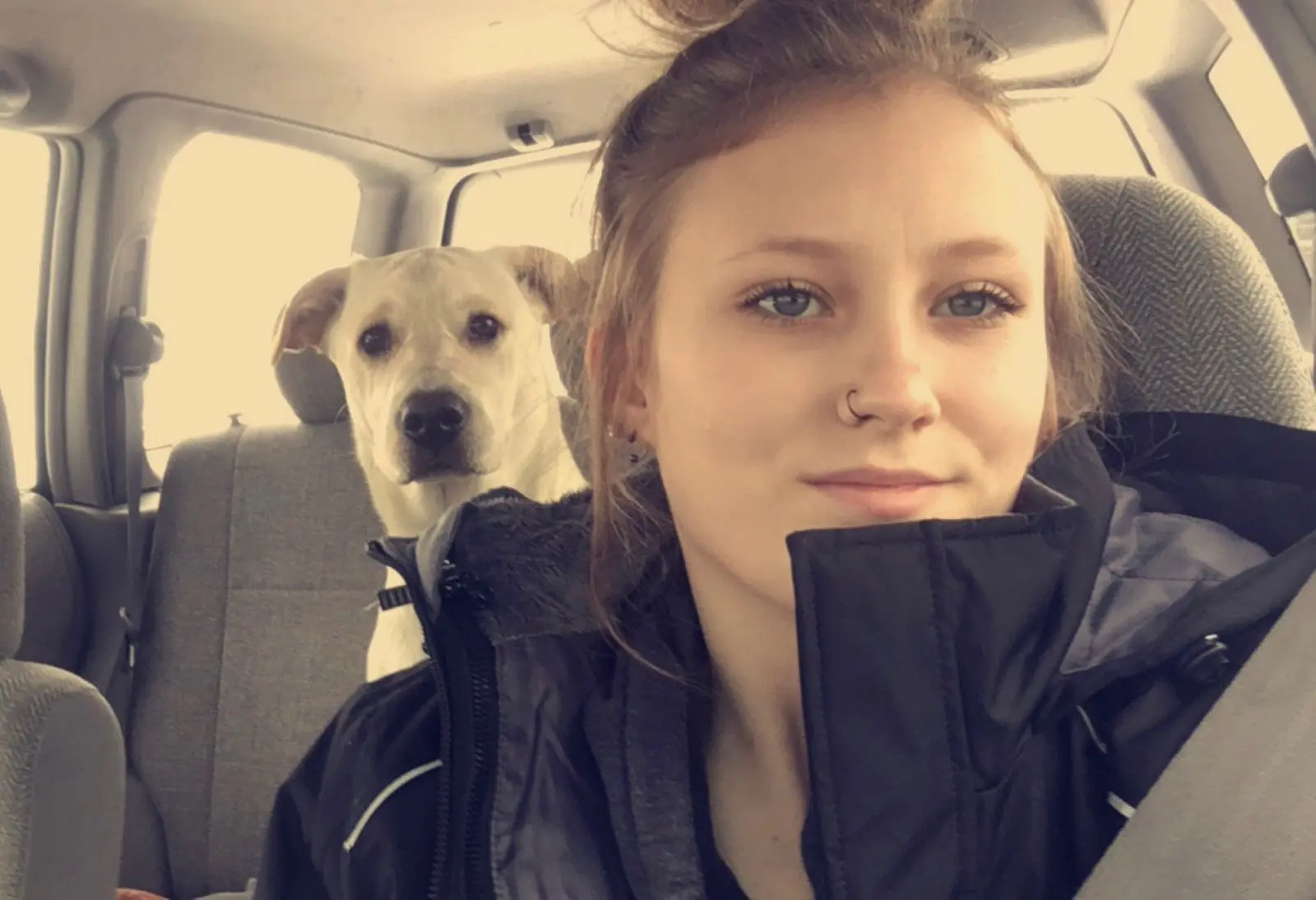 A young woman takes a selfie in a car with a dog in the backseat.