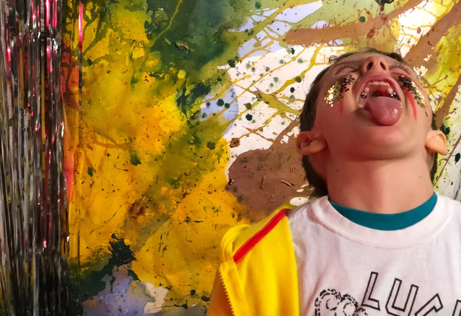A person with glitter makeup and their tongue out poses in front of a colorful painted backdrop.