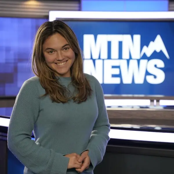 A smiling person stands in front of a news screen.